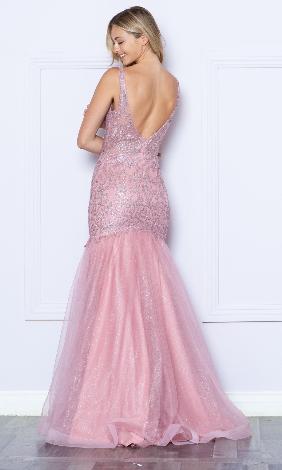 A woman faces a wall wearing the Low V-Neck Long Glitter Mermaid Prom Dress 9388, a backless, floor-length pink gown with lace, tulle details, and subtle glitter.